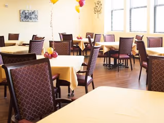 A bright dining room with multiple tables covered in yellow tablecloths and surrounded by cushioned chairs with patterned backs. There are windows letting in natural light and some balloons tied to a few chairs.