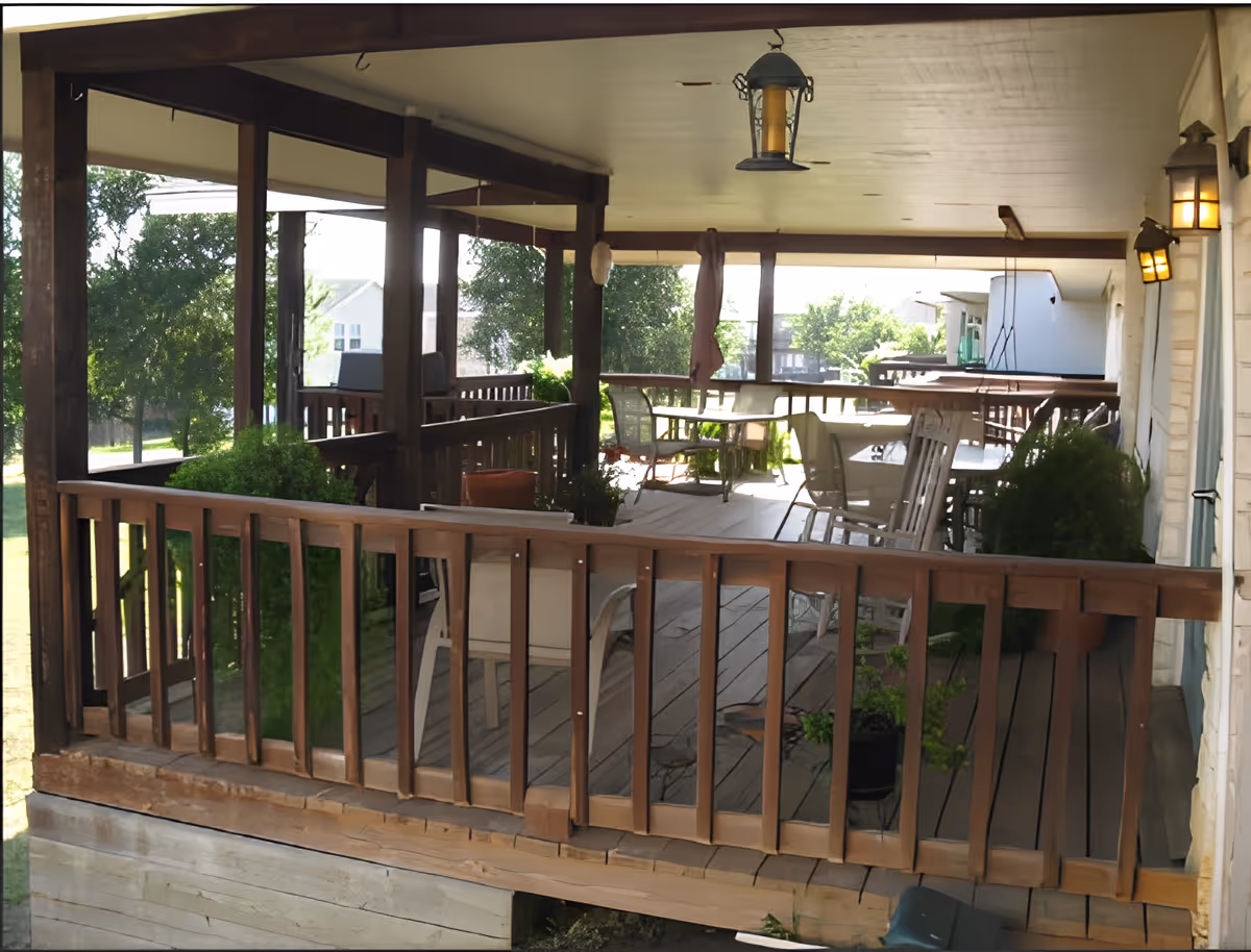 A spacious covered wooden porch with multiple tables and chairs arranged for outdoor seating. The porch is surrounded by wooden railings and has hanging lantern-style lights on the ceiling and wall. There are potted plants placed along the edges, and trees and houses are visible in the background.