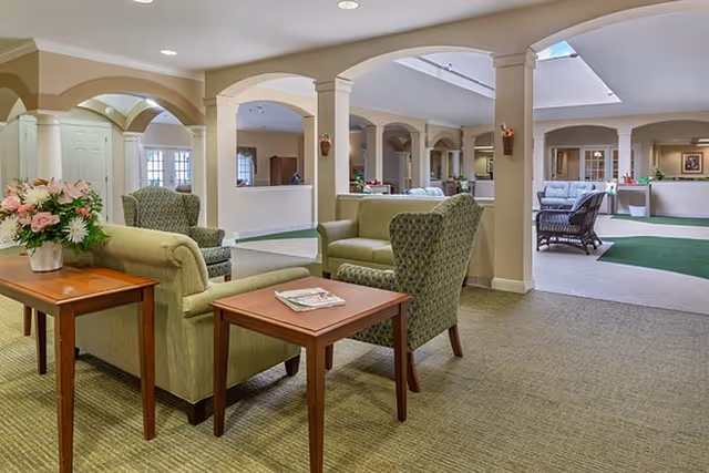 A spacious and well-lit common area in a senior living facility featuring green upholstered armchairs and sofas arranged around wooden side tables. The room has beige walls with white columns and arches, carpeted floors, and a skylight allowing natural light to brighten the space. There are floral arrangements on the tables and additional seating visible in the background.