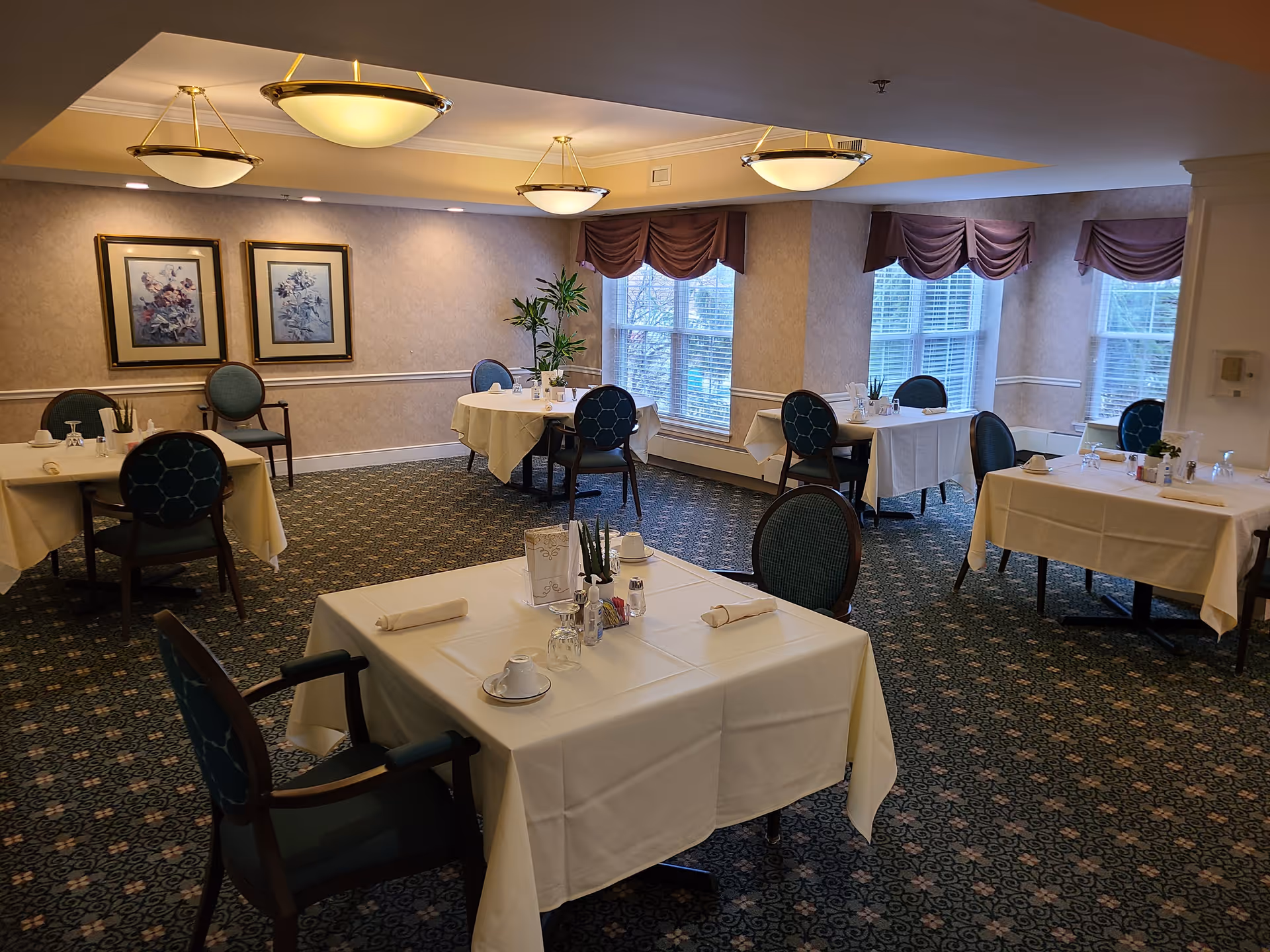A dining room with several tables covered in white tablecloths, each set with cups, napkins, and utensils. The room has patterned carpet, framed floral artwork on the walls, large windows with purple valances, and ceiling lights providing warm illumination.