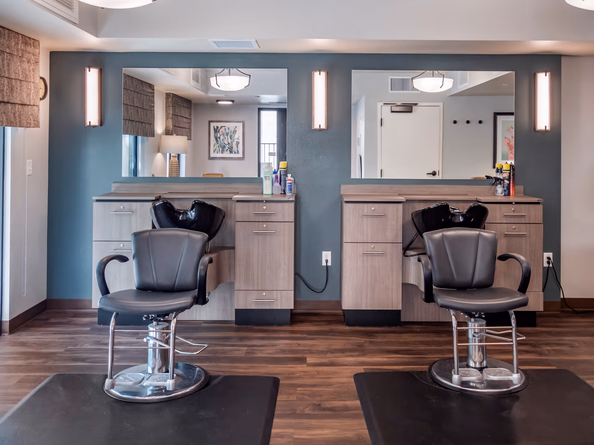 Interior view of a hair salon area with two black salon chairs in front of two large mirrors mounted on a blue-gray wall. Each station has a wooden cabinet with drawers and hair washing sinks behind the chairs. The floor is wooden, and there are wall-mounted lights beside each mirror. A window with brown Roman shades and a framed picture are visible in the background.