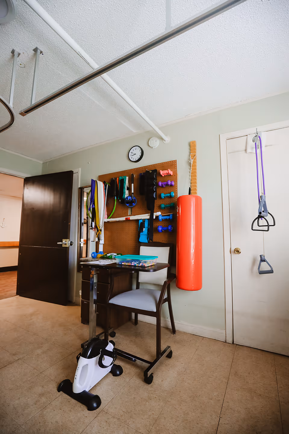 A small exercise room with a chair and a table on wheels, a mini exercise bike on the floor, and a pegboard on the wall holding various small dumbbells, resistance bands, and other exercise equipment. A red punching bag hangs on the wall next to a closed white door with purple resistance bands hanging from a hook. The room has a beige tiled floor and light green walls.