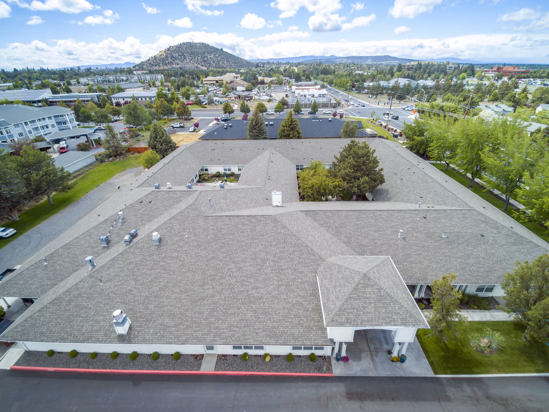 Aerial view of Awbrey Place Assisted Living and Memory Care building with a large, gray shingled roof and a covered entrance. Surrounding the building are trees, parking lots, roads, and a distant hill under a partly cloudy sky.