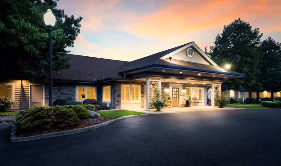 Front exterior of a single-story senior living building at dusk, showing a lit entrance portico, driveway, and landscaped grounds.