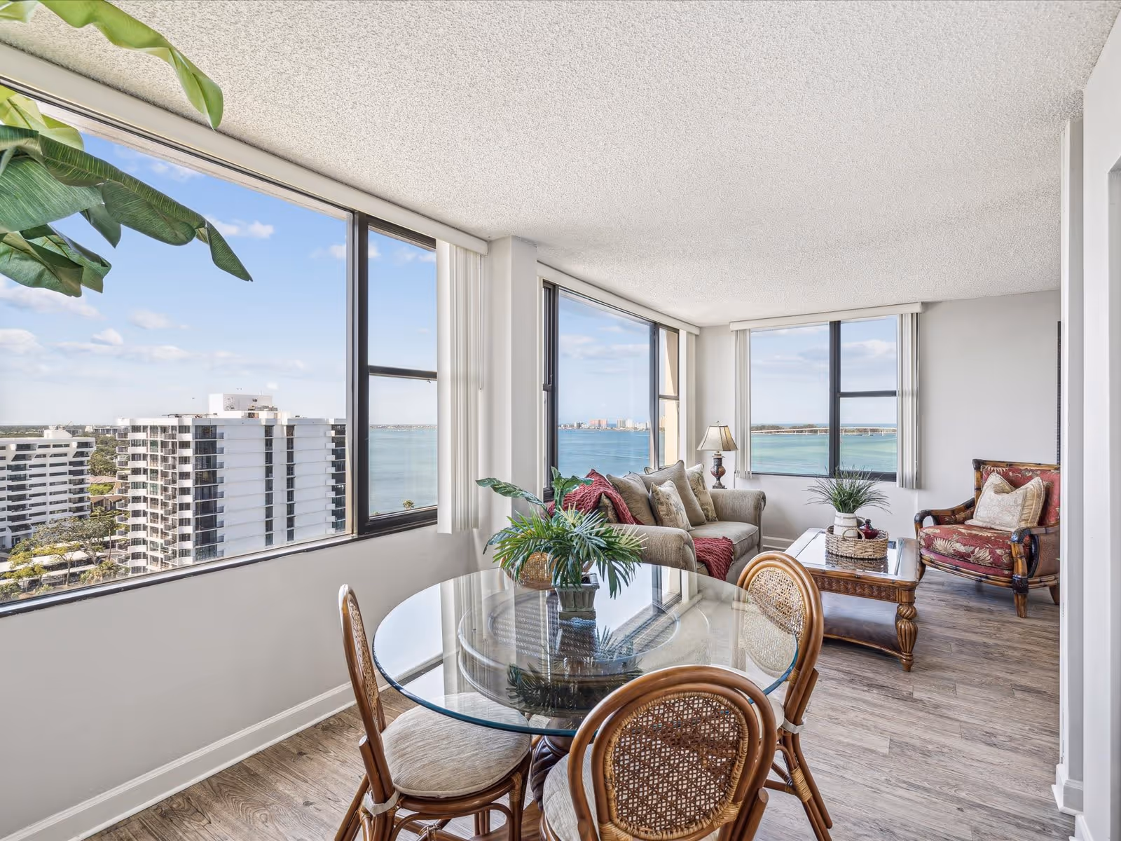 A bright living room with large windows offering a panoramic view of the ocean and nearby buildings. The room features a glass dining table with four wicker chairs, a beige sofa with cushions and a red throw, a wooden coffee table with decorative plants, and a patterned armchair. The floor is wooden, and there are vertical blinds on the windows.