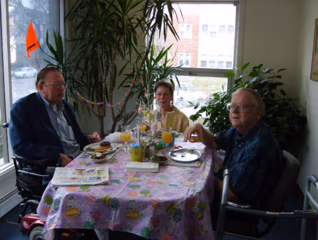 Three older adults sit around a table set with food and drinks in a bright room with large plants and windows.