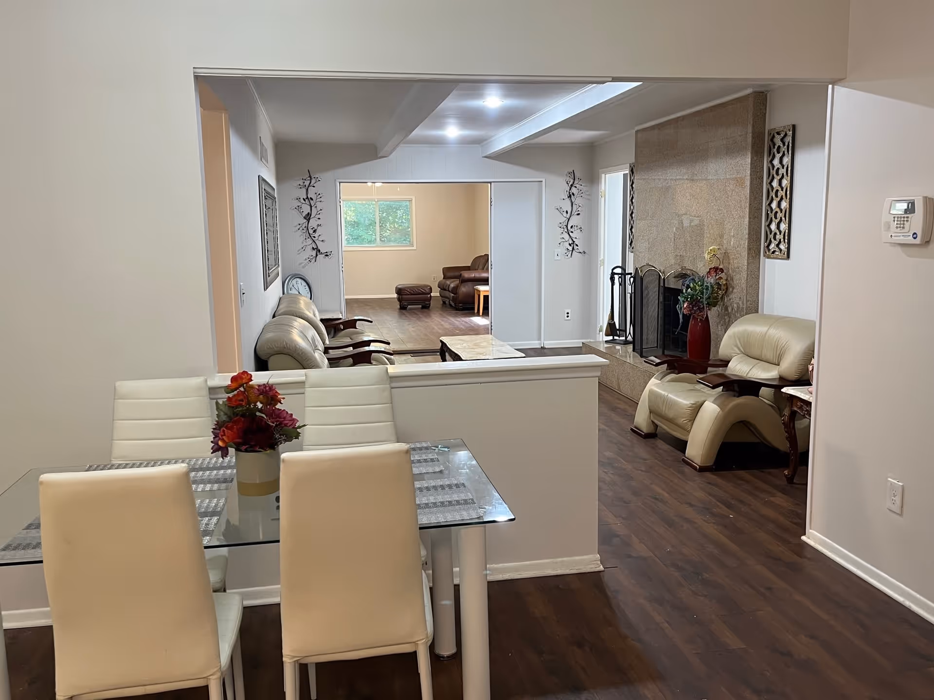 Interior view of a senior living facility showing a dining area with a glass table and white chairs in the foreground, and a living room with beige leather chairs, a fireplace, and a brown leather sofa in the background. The walls are light-colored and the floor is dark wood.
