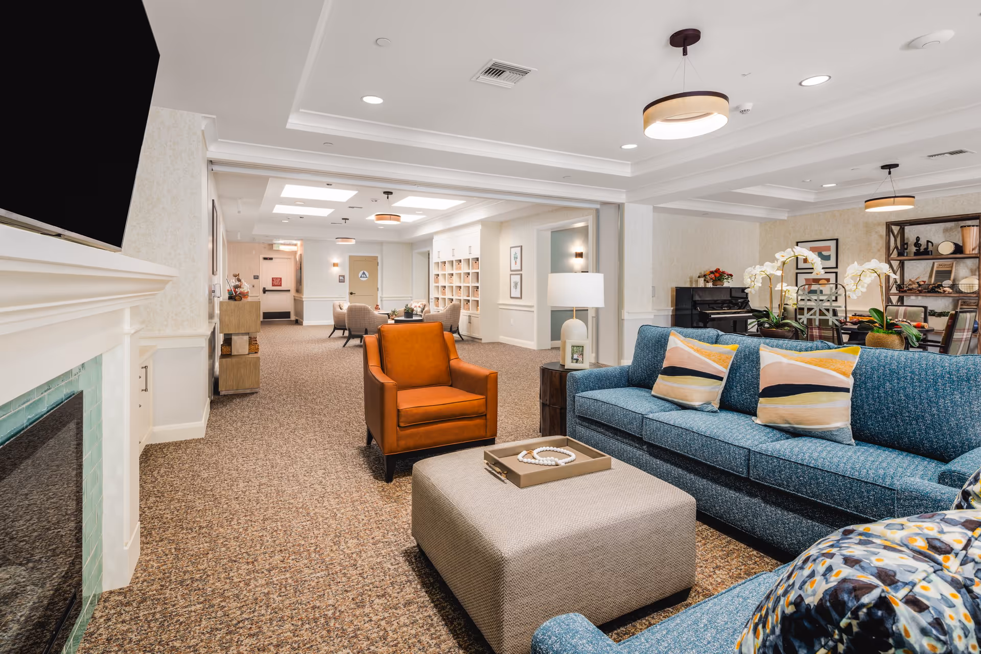 A spacious and well-lit living room area in a senior living facility featuring a blue sofa with colorful pillows, an orange armchair, a large beige ottoman with a decorative tray, a white lamp on a side table, and a fireplace with a mounted TV above it. In the background, there are additional seating areas, a piano, and decorative shelves with plants and artwork on the walls.