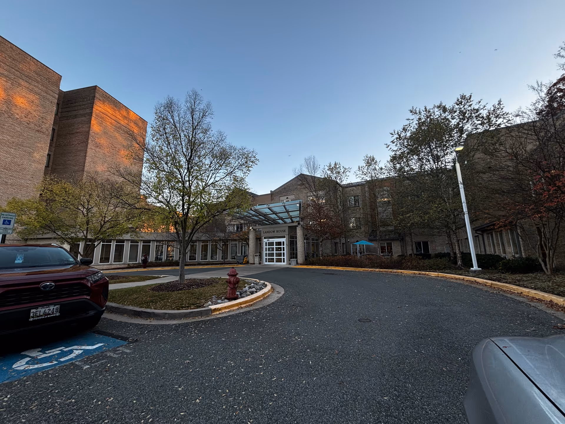 Exterior view of Landow House, a multi-story brick building with a glass entrance canopy. There are trees with sparse leaves and a curved driveway leading to the entrance. Several parked cars are visible, including one in a handicapped parking space.