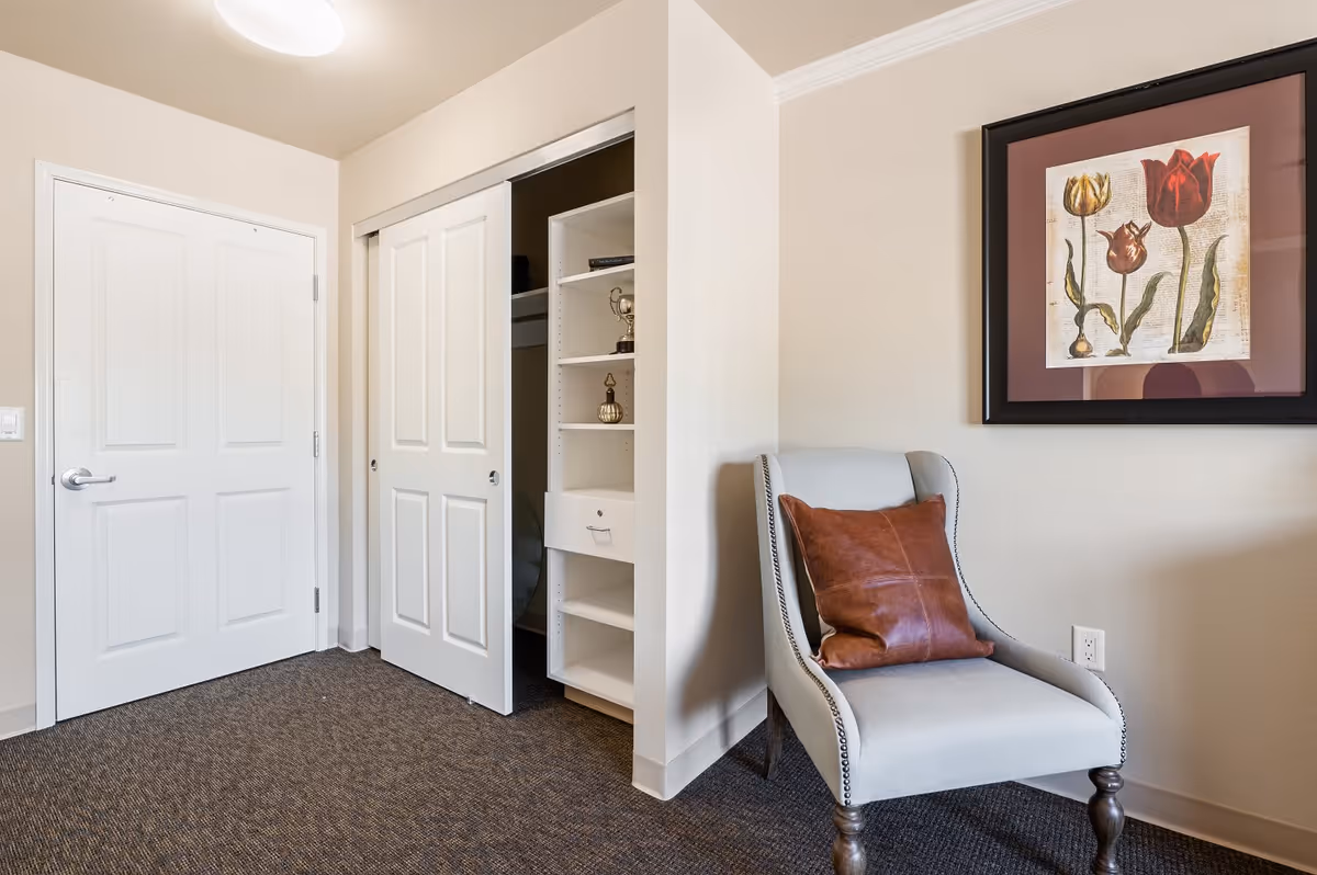 A corner of a room featuring a white door and a closet with sliding doors partially open, revealing shelves and hanging space inside. Next to the closet is a light gray upholstered chair with a brown leather pillow. Above the chair hangs a framed artwork depicting three tulips on a beige background. The floor is carpeted in a dark pattern, and the walls are painted a light beige color.
