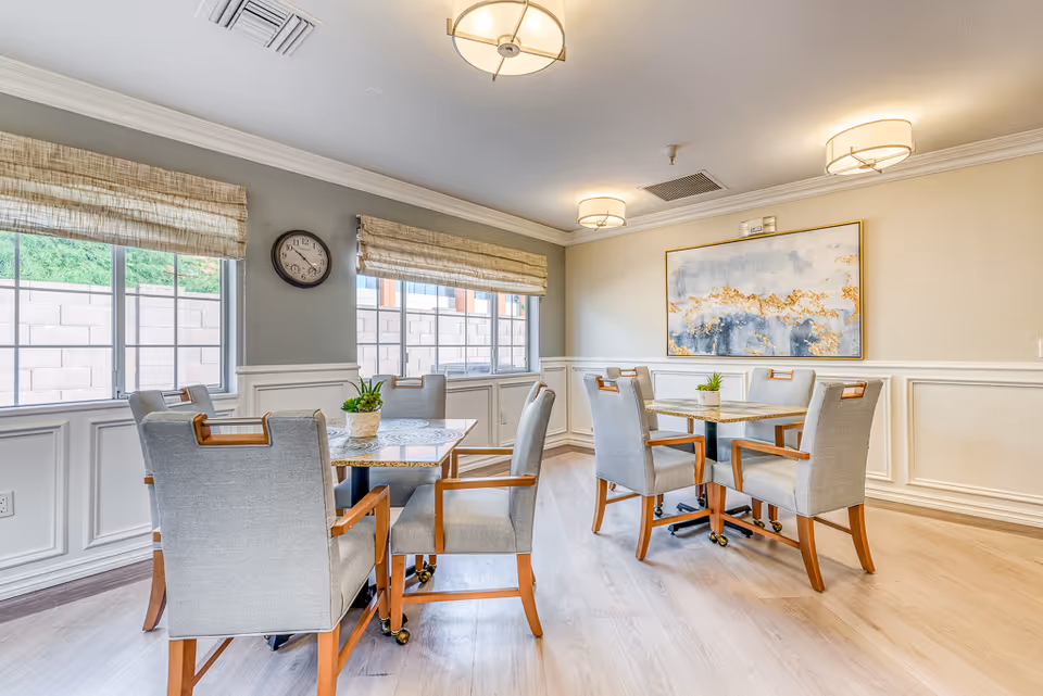 Bright common dining area with two tables and upholstered chairs, large windows, wall art and ceiling lights.