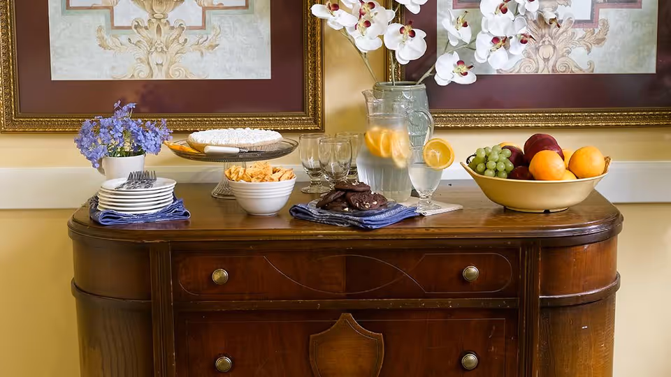 A wooden sideboard table with a bowl of fresh fruit, a pitcher of water with lemon slices, glasses, a plate of cookies, a bowl of crackers, a stack of plates with forks, and a vase with white orchids and purple flowers. Two framed decorative artworks hang on the wall behind the table.