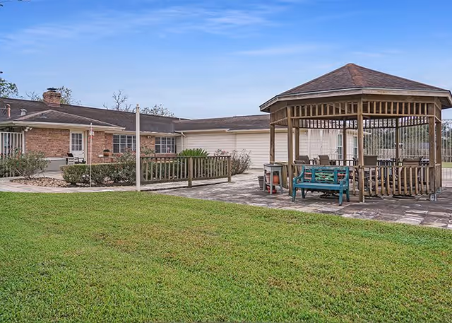 Outdoor area of a senior living facility featuring a large grassy lawn, a wooden gazebo with seating, and a single-story brick and siding building in the background under a clear blue sky.