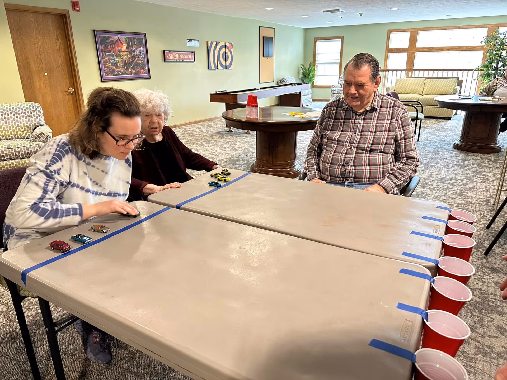Three people sitting around a table in a common area playing a game with small toy cars and red plastic cups taped to the edge of the table. The room has green walls, carpeted floor, and large windows letting in natural light. There are couches, a shuffleboard table, and framed artwork on the walls.