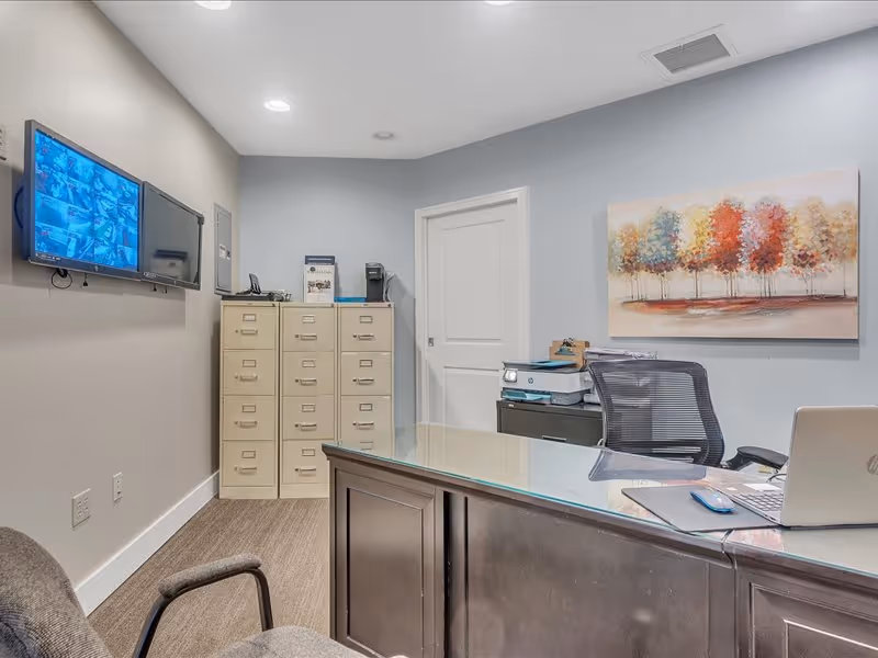 Small administrative office with a desk, filing cabinets, a wall-mounted monitor, and colorful tree artwork.