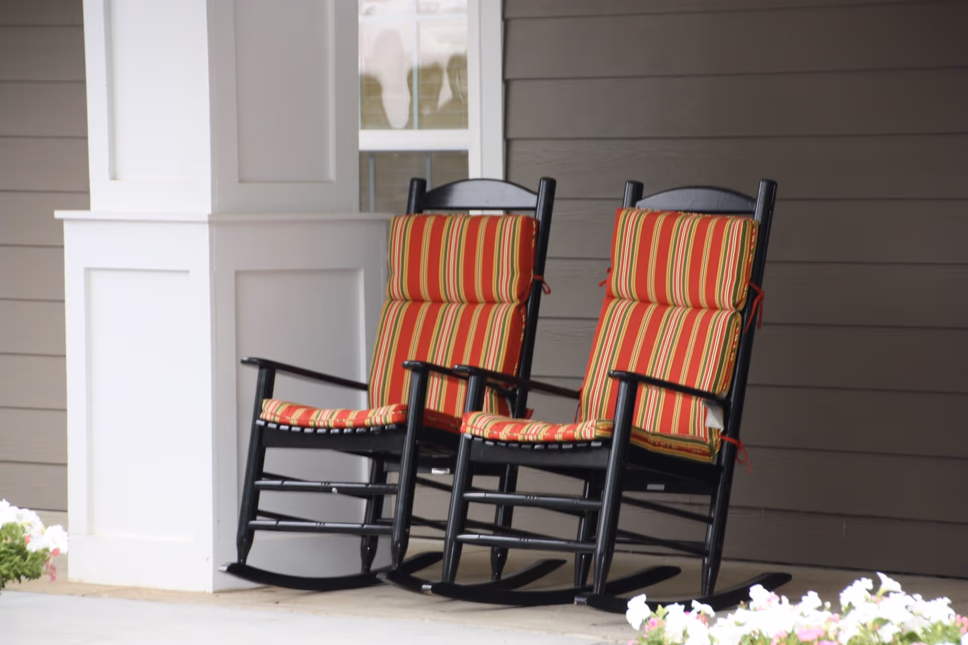 Two black wooden rocking chairs with red and yellow striped cushions placed on a covered porch with brown siding and white trim, with flowers visible in the foreground.