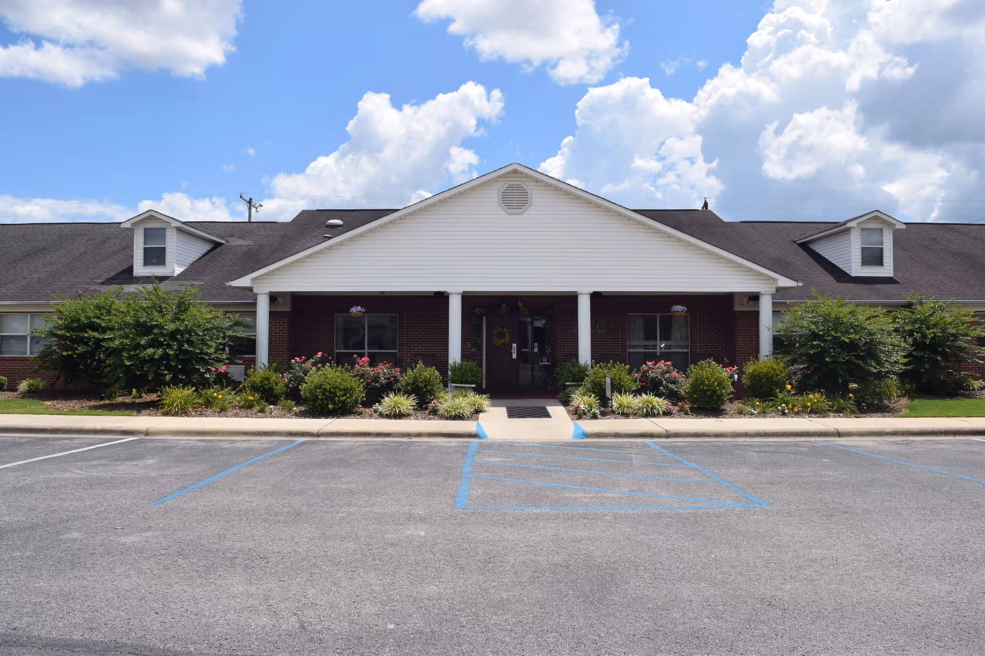 Front exterior view of Brentwood Retirement Community building with a white gabled roof, brick walls, two dormer windows, a covered entrance supported by white columns, and landscaped bushes and flowers in front. The sky is partly cloudy with blue patches.