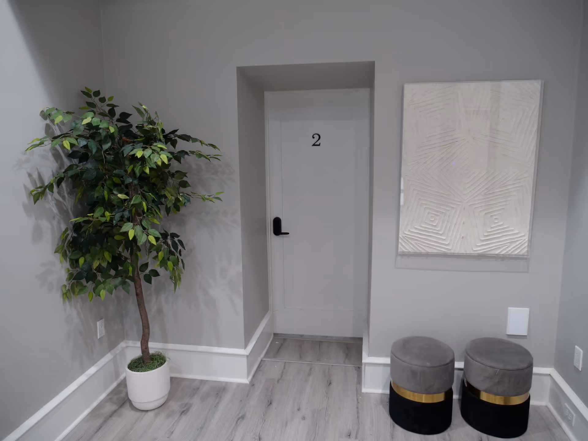 A hallway corner with a white door labeled number 2, a potted artificial tree on the left, two round gray stools with gold accents on the right, and a textured white wall art piece above the stools. The walls and floor are light gray.