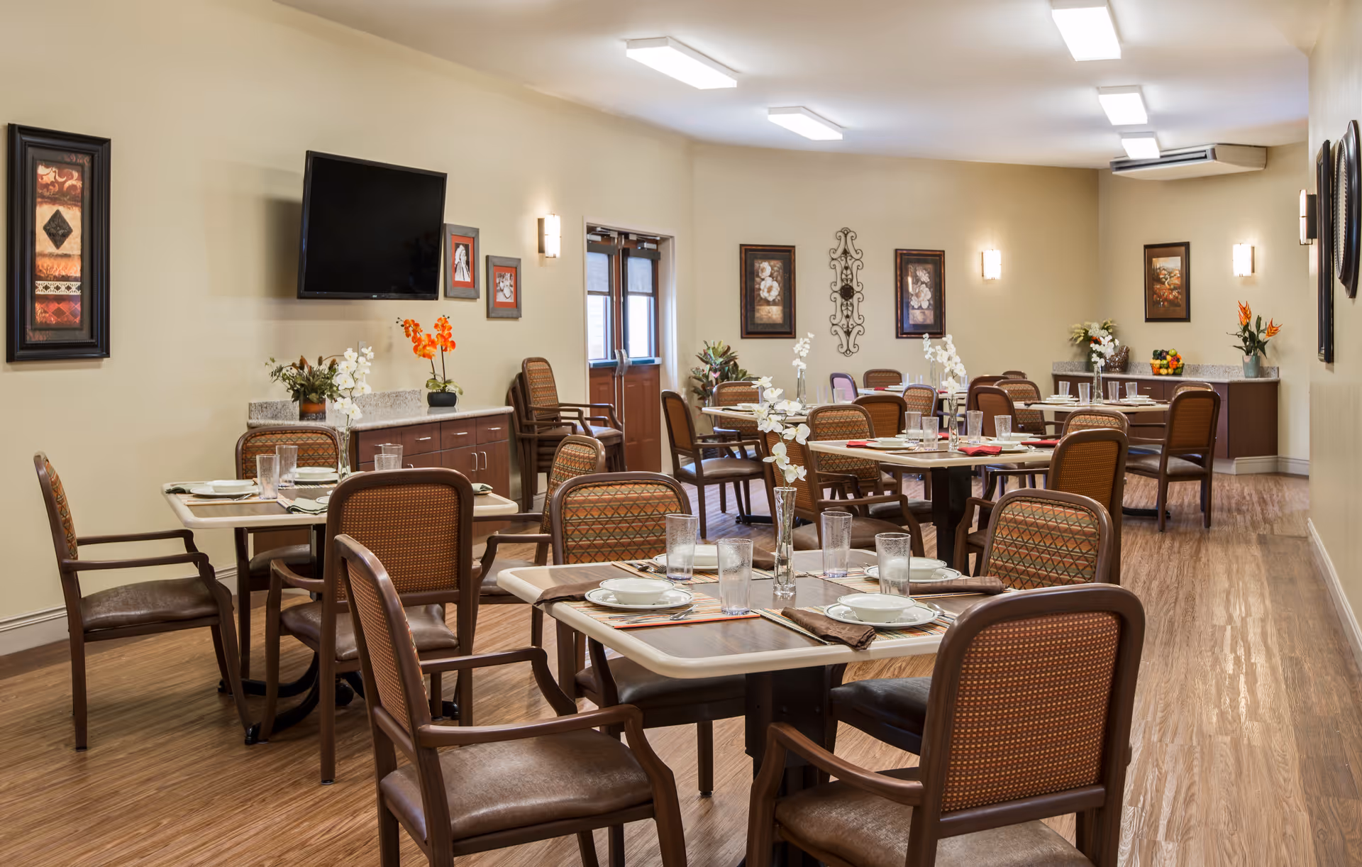 A well-lit dining room in a senior living facility with multiple tables set with plates, glasses, and napkins. The room features wooden chairs with cushioned seats, decorative wall art, a flat-screen TV mounted on the wall, and floral arrangements on the tables and sideboards.
