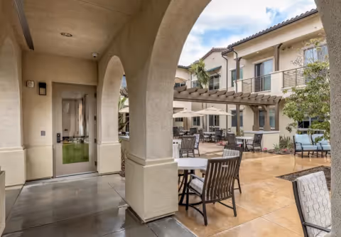 Covered archway opening onto a courtyard with outdoor dining tables, chairs, a pergola, and a two-story building.