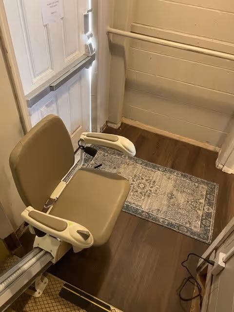 A beige stairlift chair installed on a stairway with wooden flooring and a patterned rug at the bottom. The stairlift is positioned near a white door with a horizontal push bar and a sign taped to it. The walls are light-colored with a handrail on one side.