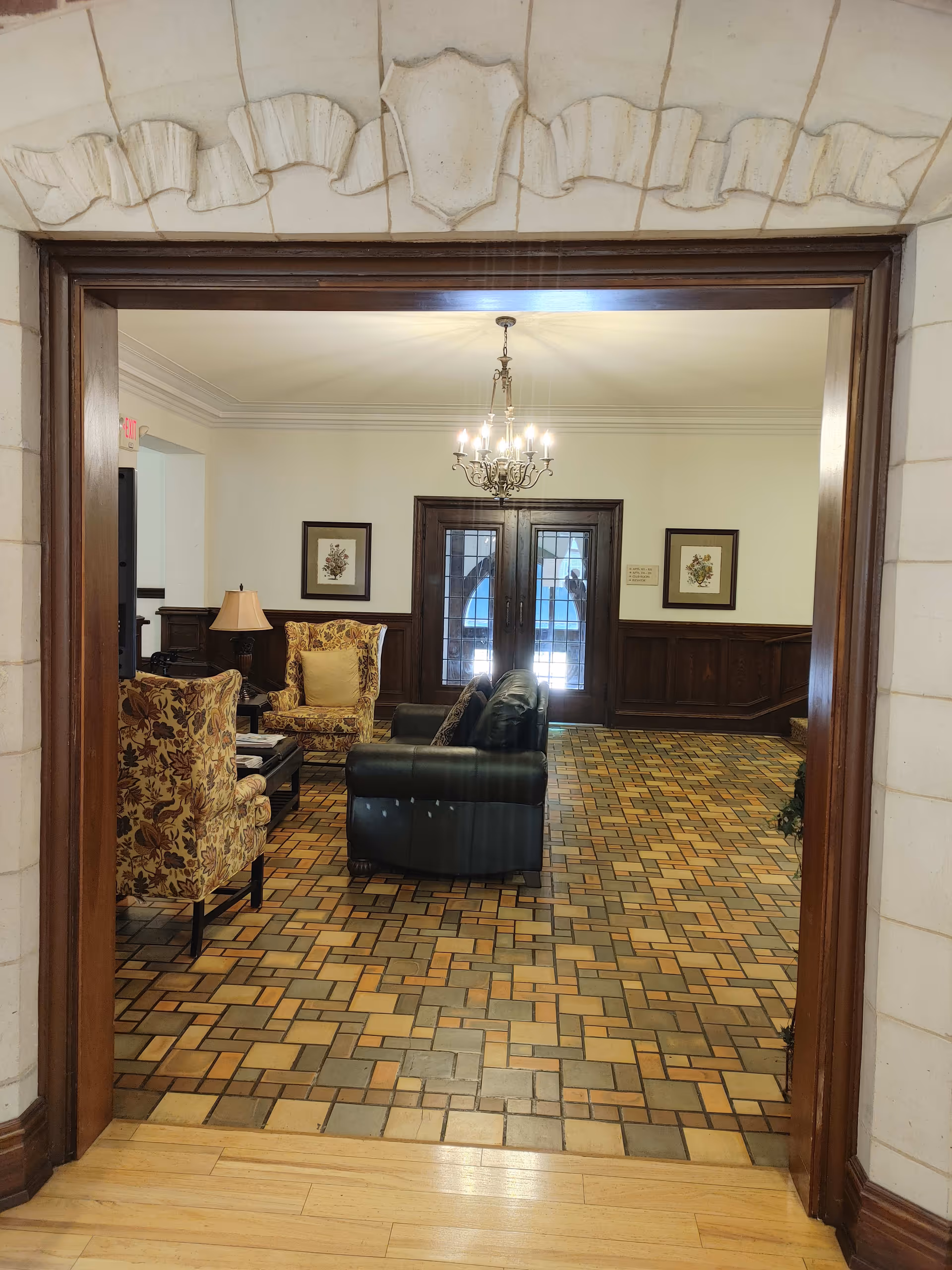 View through a doorway into a sitting area with patterned tile flooring, a black leather sofa, two floral upholstered armchairs, a wooden coffee table, a table lamp, and a chandelier hanging from the ceiling. The back wall features double glass doors and two framed floral artworks.
