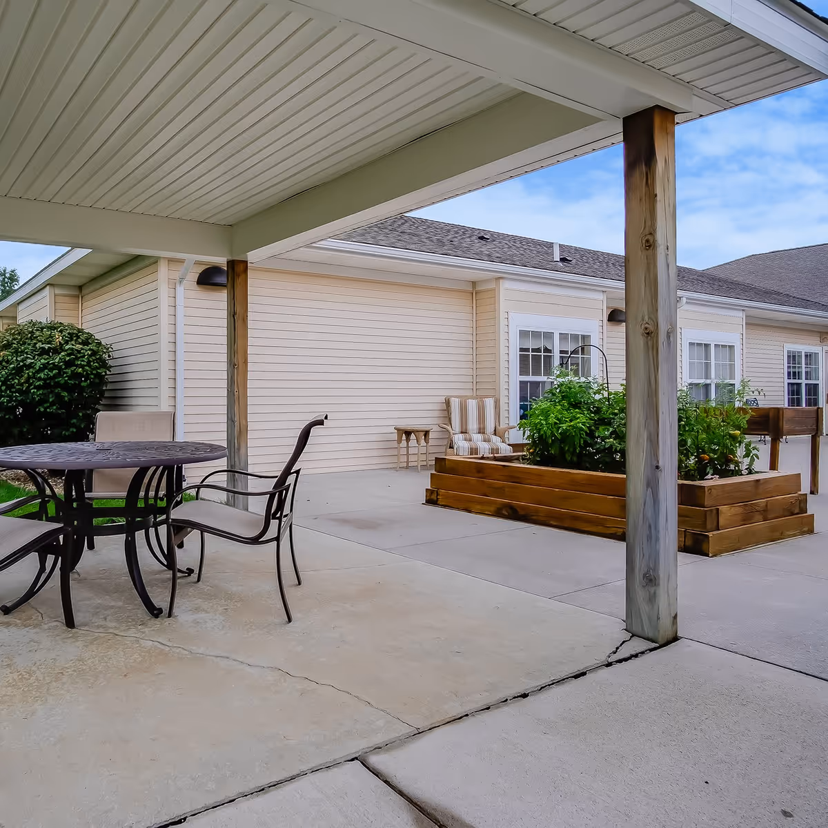 Covered outdoor patio area with a round metal table and chairs, a cushioned armchair, wooden planter boxes with green plants, and beige siding building walls under a blue sky with some clouds.