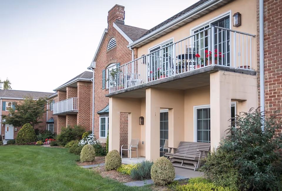 Exterior view of a senior living facility building with brick and beige walls, featuring balconies with patio furniture and flower pots. There is a well-maintained lawn and landscaped bushes in front of the building.