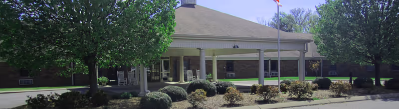 Front exterior view of Village Manor Assisted Living building with a covered entrance supported by white columns, surrounded by green trees and landscaped bushes under a clear sky.