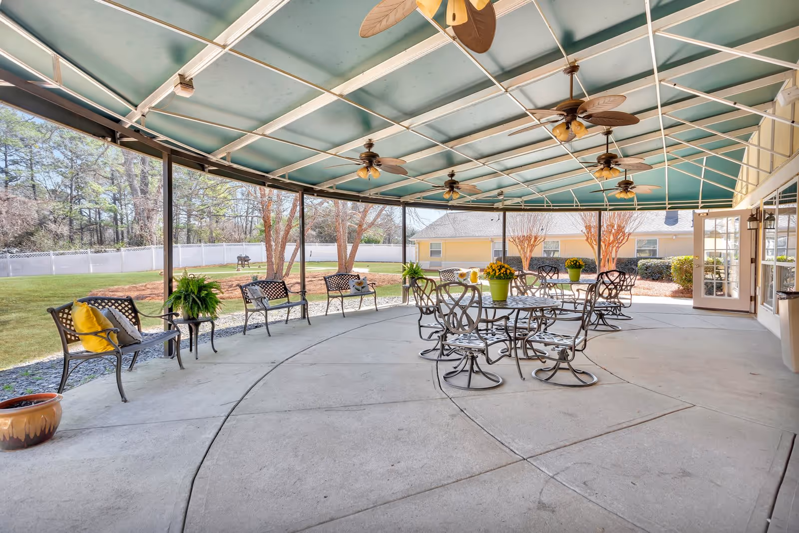 Covered outdoor patio area with metal tables and chairs, ceiling fans, and benches with cushions. The patio overlooks a grassy yard with trees and a white fence, and a yellow building is visible in the background.