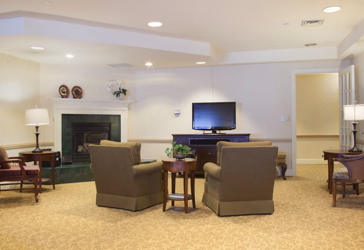 Comfortable lounge with two upholstered armchairs facing a TV, a fireplace, side tables, and lamps in a neutral-decor room.