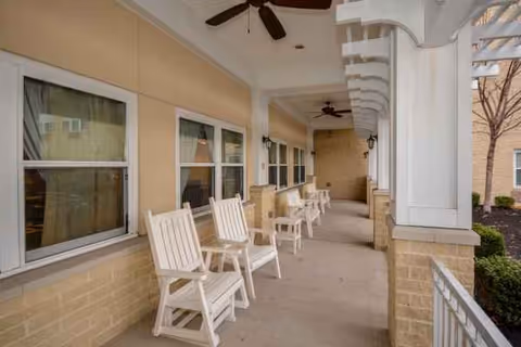 Covered outdoor porch with white rocking chairs along a beige facility exterior.