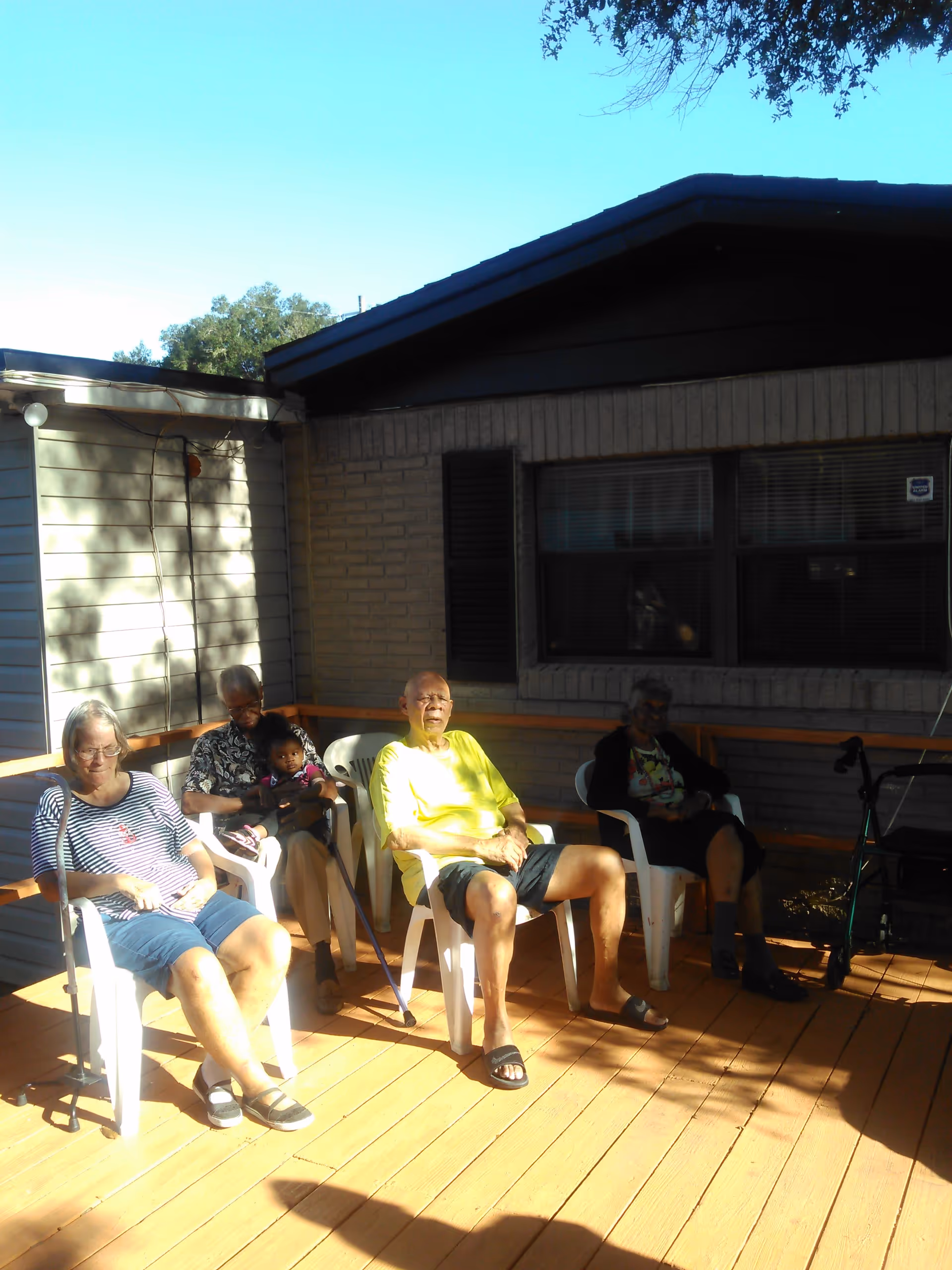 Four elderly people and one child sitting on white plastic chairs on a wooden deck outside a brick building. The elderly individuals appear relaxed, enjoying the sunny weather, with one using a cane and another having a walker nearby.