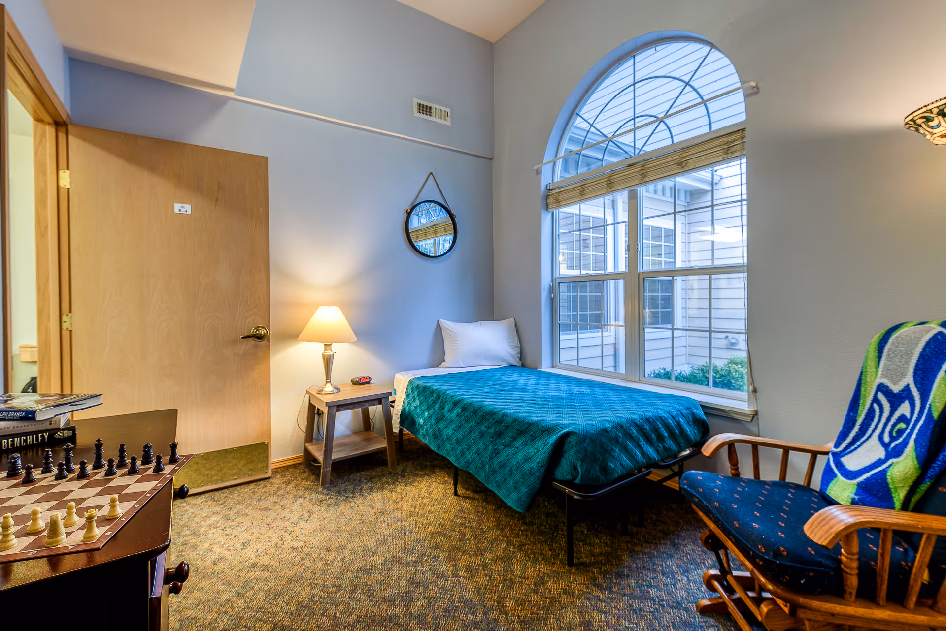 A small bedroom with a single bed covered with a teal blanket, a wooden nightstand with a lamp and digital clock, a round mirror hanging on the wall, a large arched window with blinds, a wooden rocking chair with a colorful blanket draped over it, and a table with a chessboard and books.