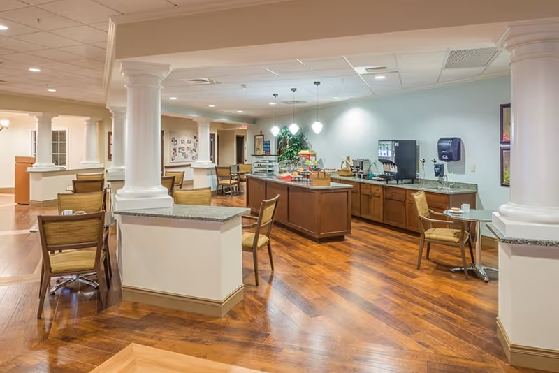 A spacious dining area in an assisted living facility with wooden floors, several tables and chairs, white columns, and a counter with a coffee machine and snacks. The room is well-lit with ceiling lights and pendant lamps above the counter.