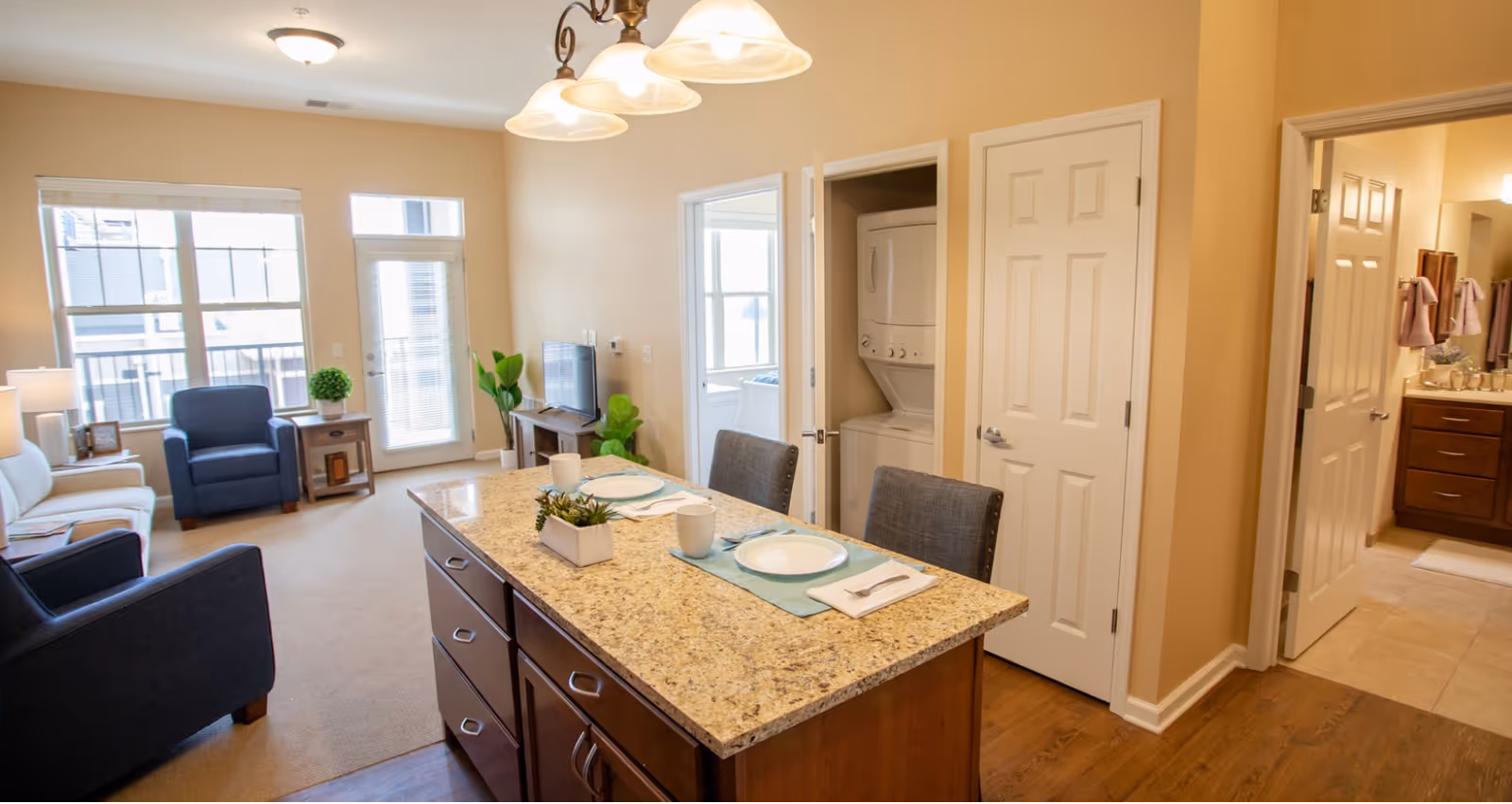 Interior view of a senior living apartment featuring a kitchen island with two place settings, a living area with a white sofa and blue armchairs, a TV on a stand, a door leading to a balcony, a laundry closet with stacked washer and dryer, and a bathroom visible through an open door.