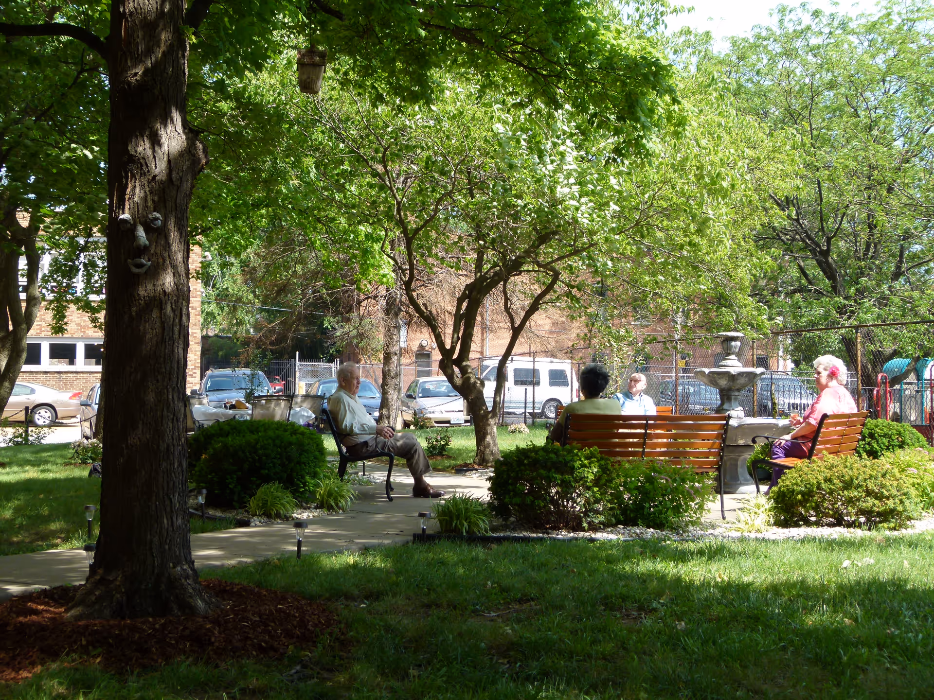 An outdoor garden area with four elderly people sitting on benches arranged around a stone fountain. The area is shaded by trees and surrounded by green bushes and grass. A tree with a face decoration is visible in the foreground, and parked cars and a brick building are seen in the background.
