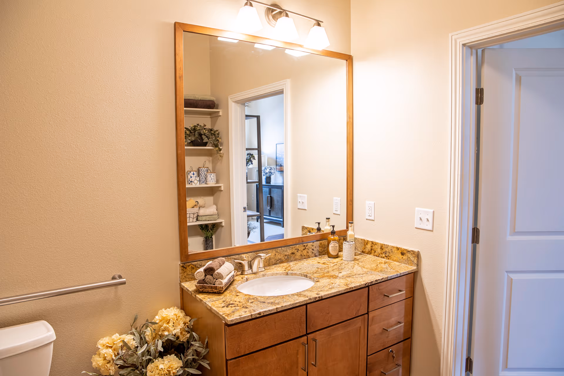 A bathroom with a granite countertop vanity featuring a sink, soap dispensers, and neatly rolled towels. There is a large wooden framed mirror above the vanity reflecting an adjacent room with shelves and decorative items. A toilet is partially visible on the left side, and a white door is open on the right side. The walls are painted beige and there is a towel bar mounted on the wall.