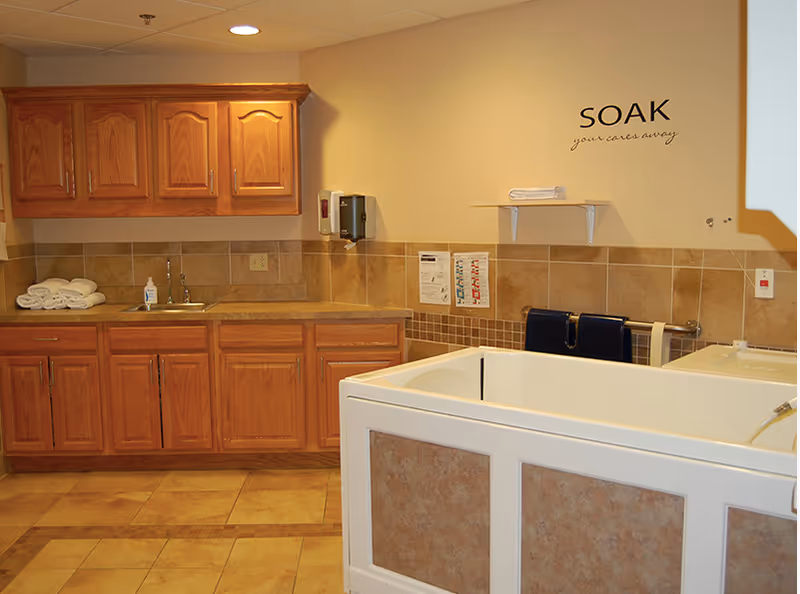 Interior view of a bathing area in a senior living facility featuring a large white walk-in bathtub with tiled panels, wooden cabinets with a countertop and sink, folded towels, soap dispenser, and a wall decal that reads 'SOAK your cares away'.