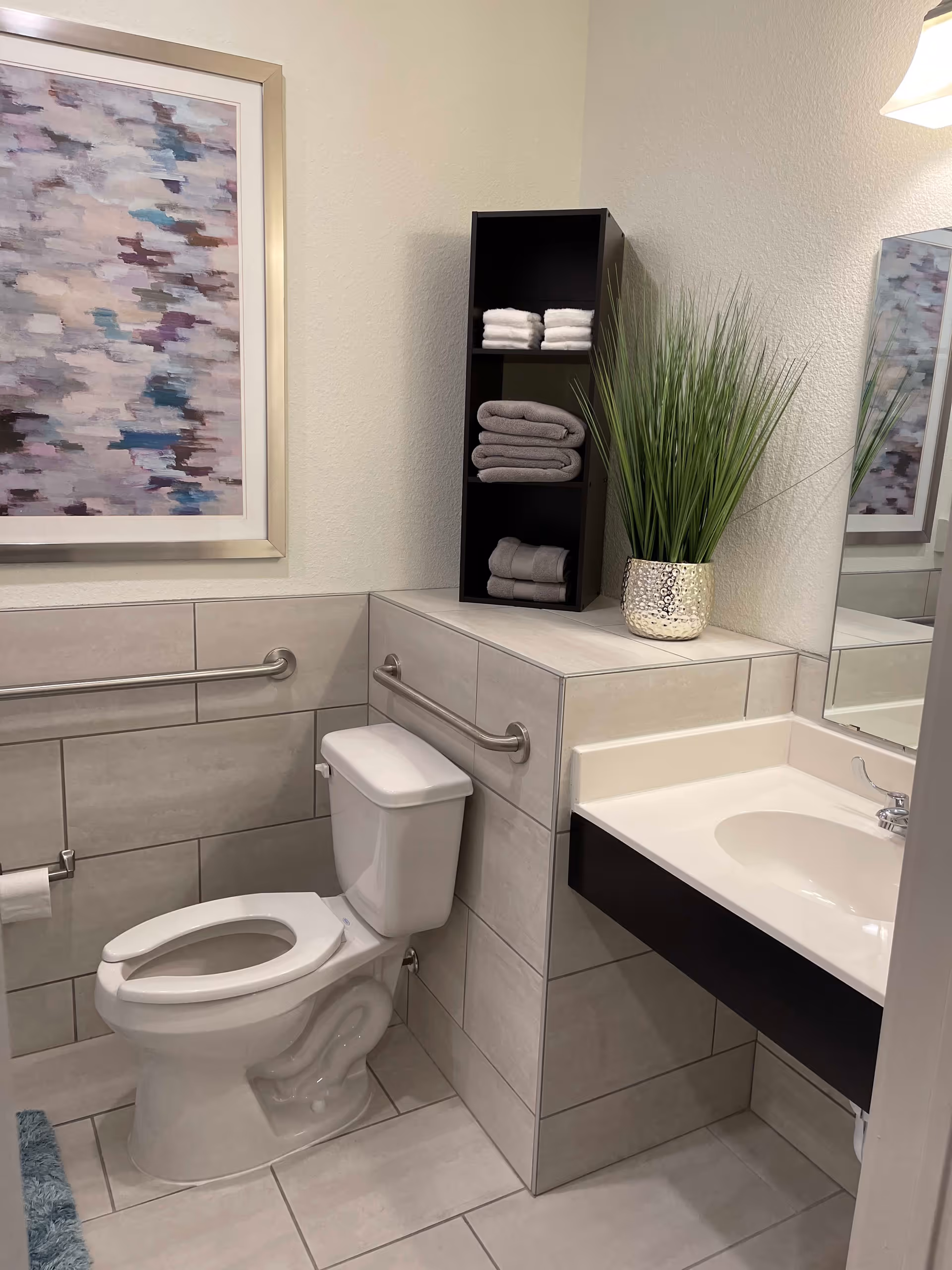 A clean and modern bathroom with a white toilet, a white sink with a silver faucet, and a black shelving unit holding neatly folded white and gray towels. There is a decorative plant in a silver pot on the counter next to the sink, a large mirror above the sink, and a framed abstract painting on the wall. The walls and floor are tiled in light gray, and there are metal grab bars on the walls near the toilet.
