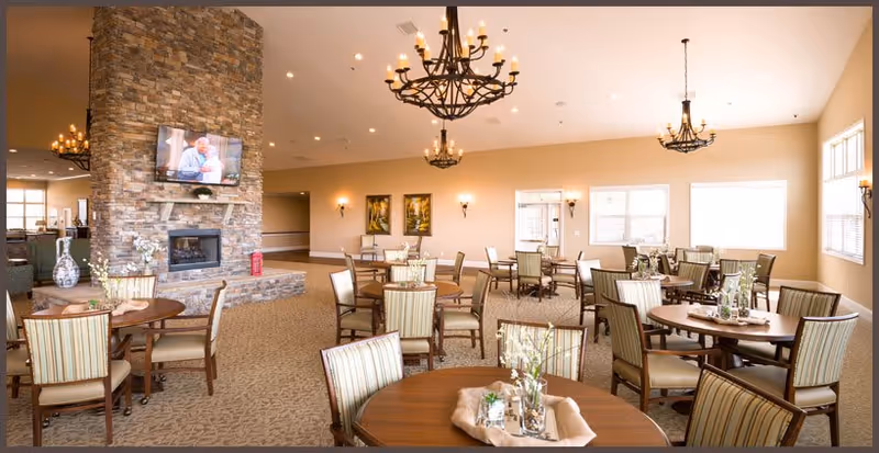 Spacious dining room with round tables and upholstered chairs, chandeliers, and a stone fireplace with a mounted TV.