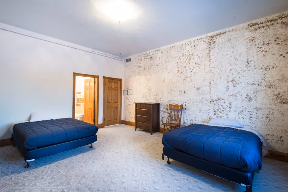A senior living facility bedroom with two single beds covered in dark blue comforters, a wooden dresser, a wooden chair, and a door leading to a bathroom. The room has a textured wall and light-colored carpet flooring.