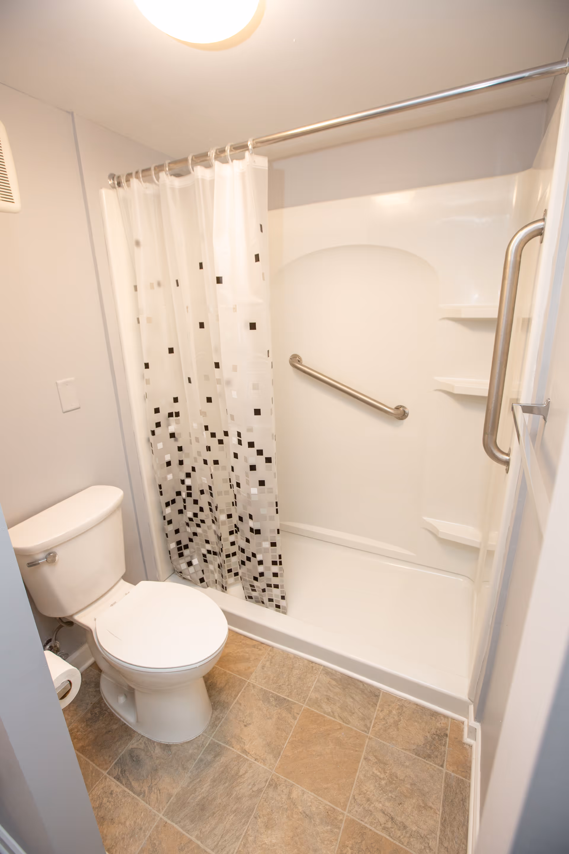 A small bathroom with a white toilet and a shower area with a white curtain featuring a black and gray square pattern. The shower has built-in shelves and a metal grab bar. The floor is covered with beige and brown tiles.