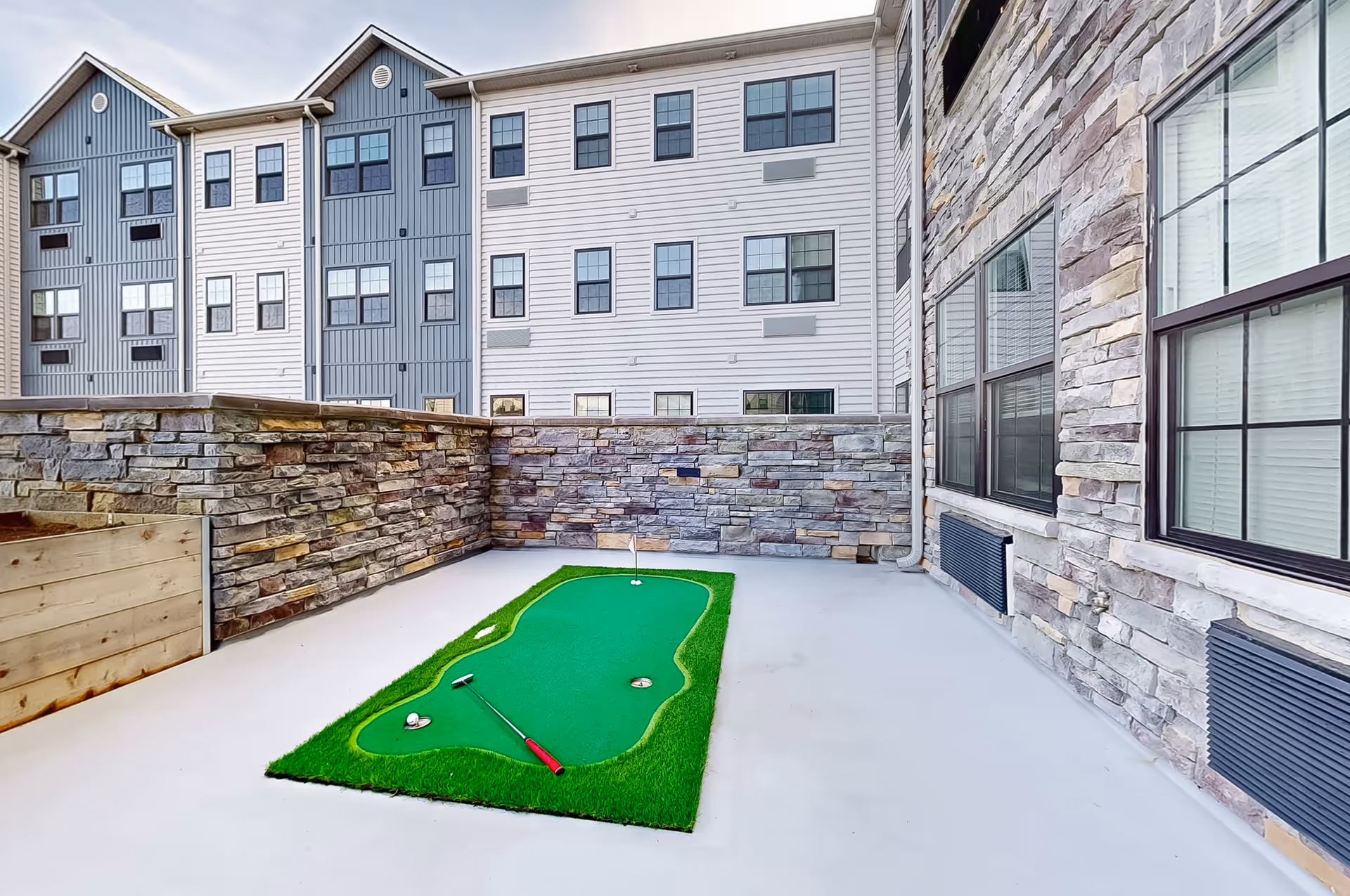 Outdoor patio area with stone walls and a small green putting mat with a golf ball and putter on a concrete floor, surrounded by multi-story residential buildings with windows.