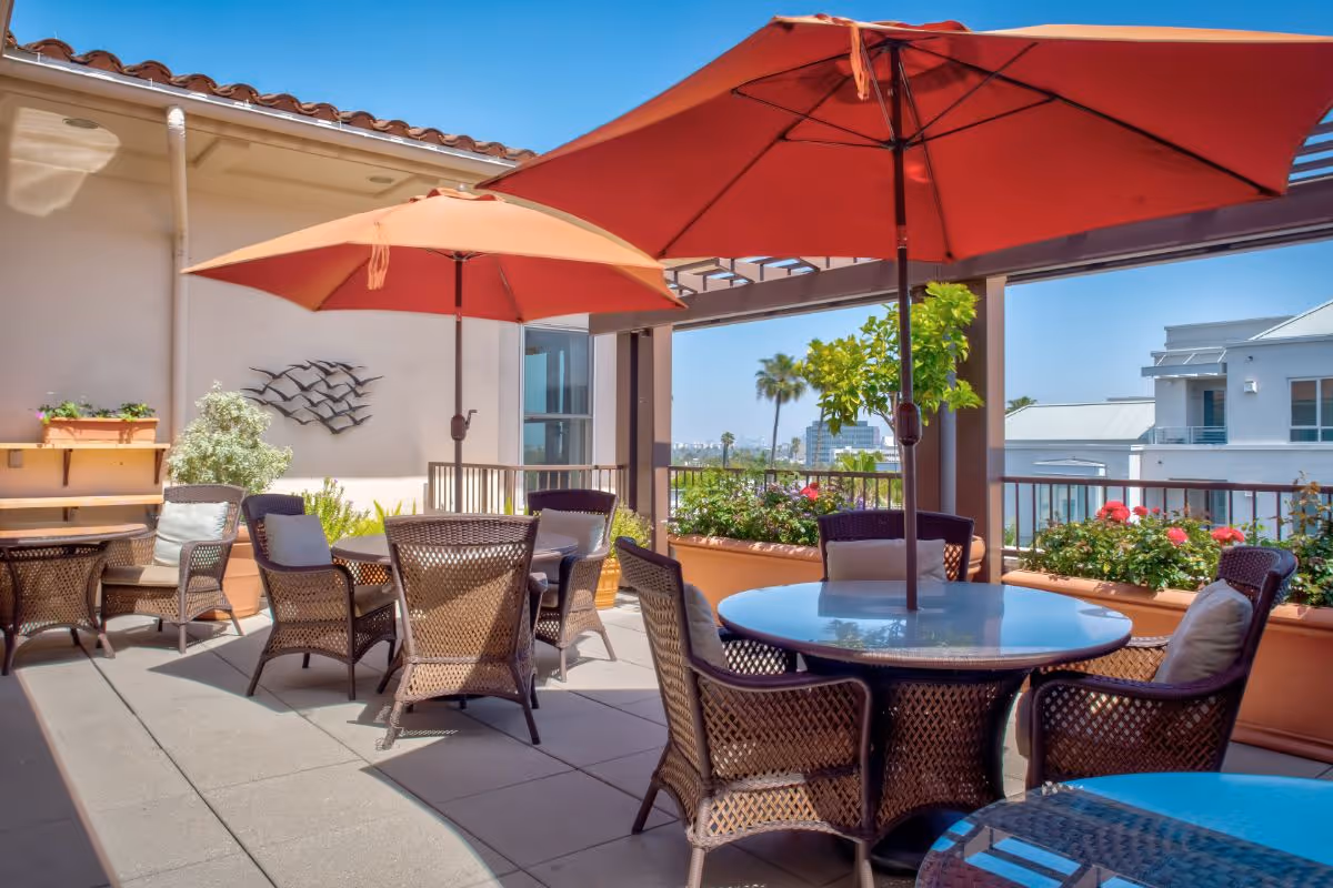 Sunny rooftop patio with wicker tables and chairs under red umbrellas and potted plants.