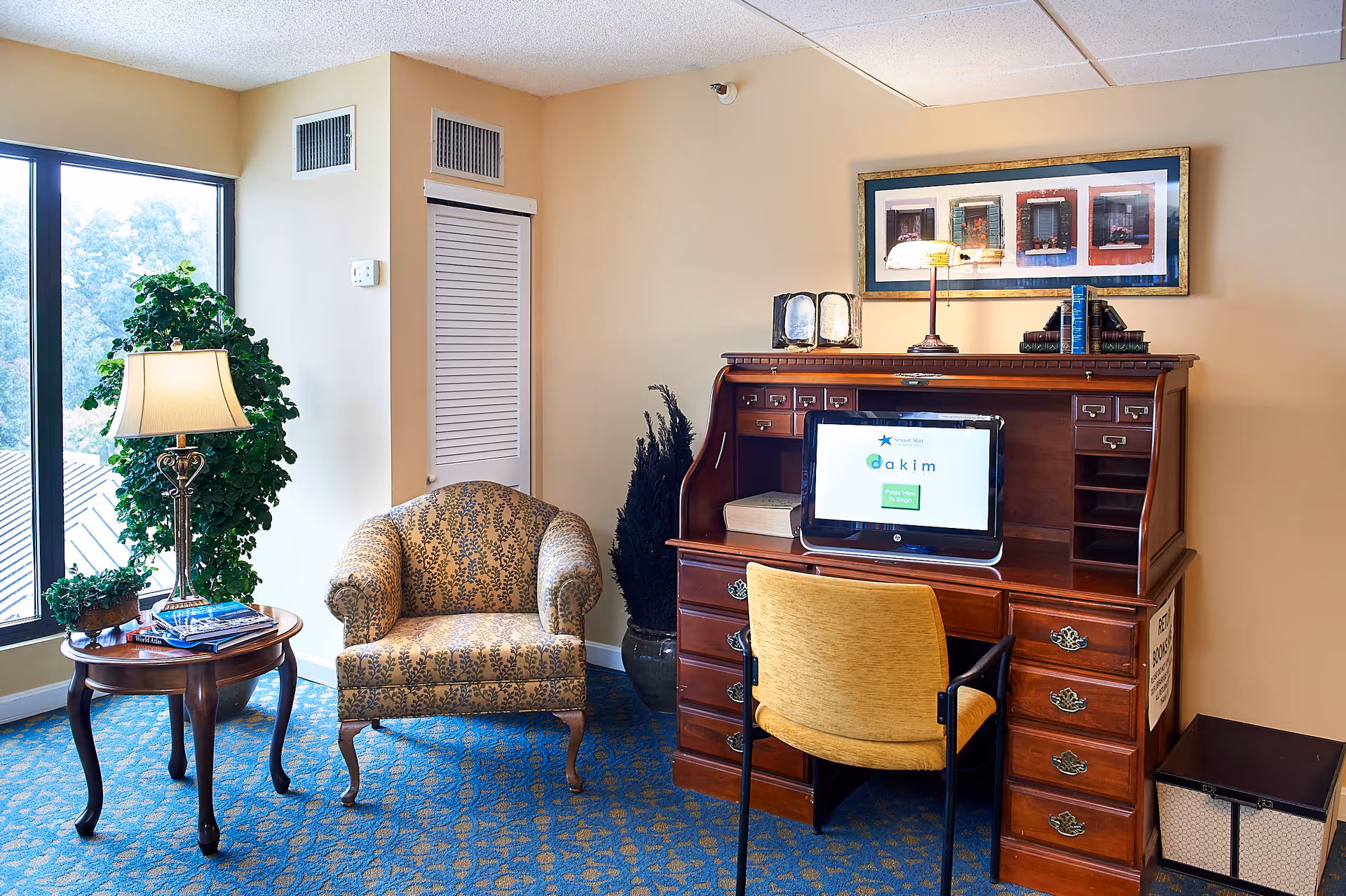 A cozy corner in a senior living facility featuring a wooden roll-top desk with a computer on it, a yellow upholstered chair, a patterned armchair, a round wooden side table with a lamp and magazines, and a large window with a view of trees outside. The room has beige walls and blue patterned carpet.