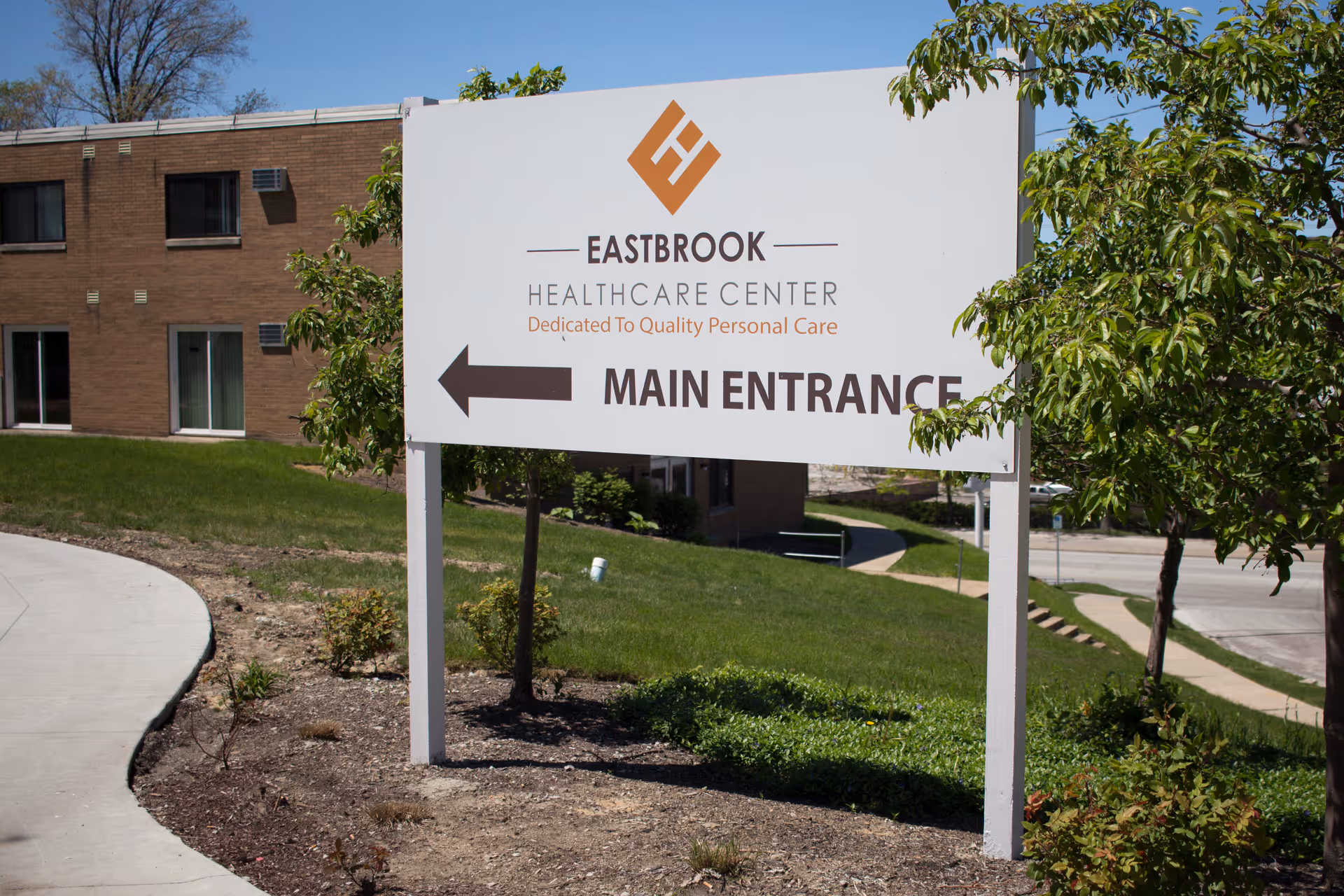 Outdoor view of a sign for Eastbrook Healthcare Center with an arrow pointing left indicating the main entrance. The sign is surrounded by grass, small plants, and trees, with a building and sidewalk visible in the background under a clear blue sky.