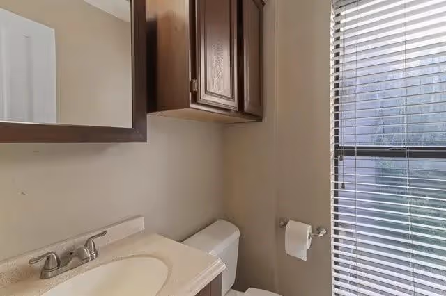A small bathroom with a white sink and countertop, a silver faucet, a wooden cabinet above the toilet, a toilet with a roll of toilet paper on a holder, and a window with blinds partially open letting in natural light.