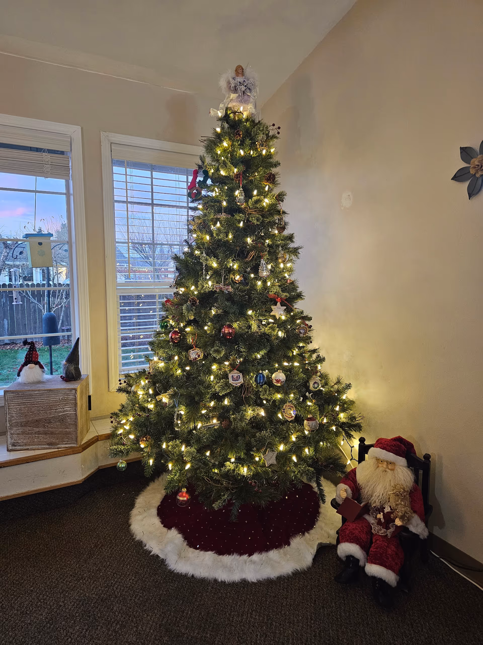 A decorated Christmas tree with white lights and various ornaments stands in a corner near two windows with white blinds. The tree has a white and red tree skirt at the base. To the right of the tree, there is a Santa Claus figure sitting in a small black chair. On the left, a wooden box with two small gnome decorations sits on a window ledge. The room has beige walls and dark carpeted flooring.