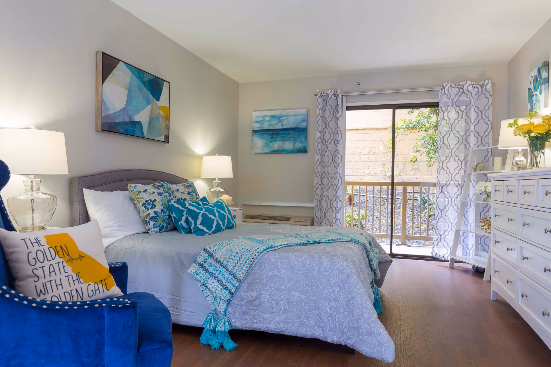 Well-lit senior bedroom with a made bed, patterned pillows and throw, a blue armchair, white dresser, and a sliding glass door leading outside.